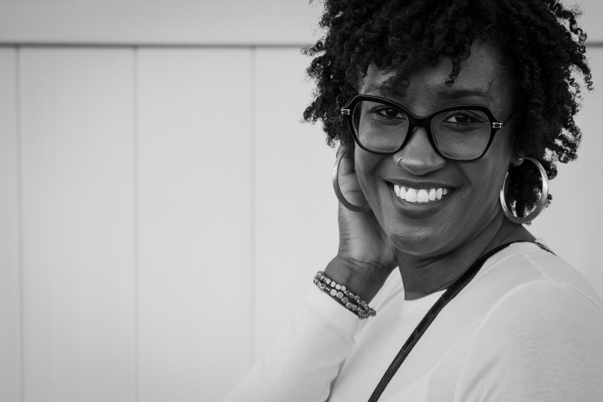 Black and white portrait of a smiling woman with curly hair and eyeglasses indoors.
