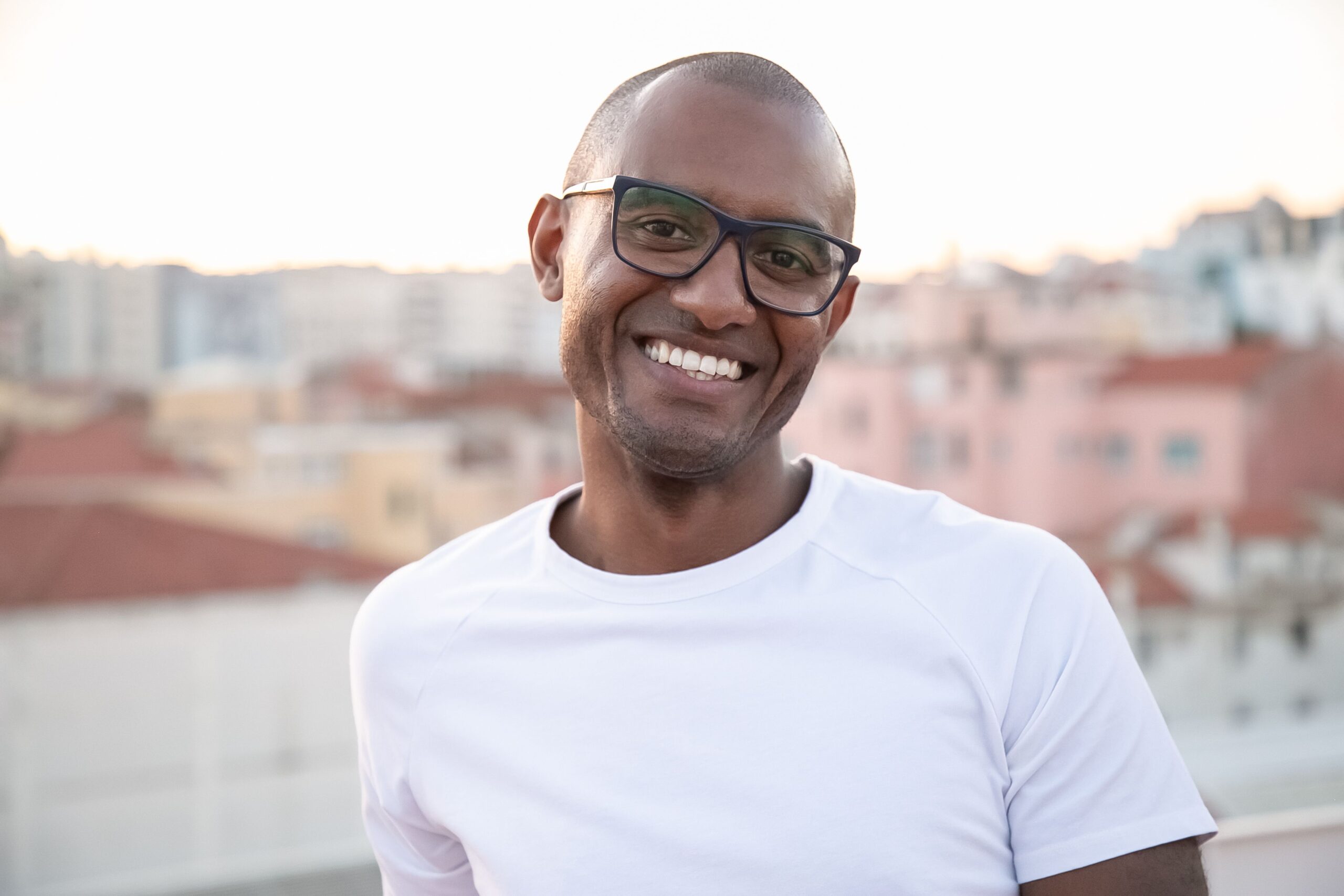 A joyful portrait of a man in a white shirt and eyeglasses smiling against an urban backdrop in Portugal.
